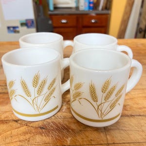 Peut inclure: Quatre tasses en céramique blanche avec des motifs dorés de tiges de blé. Chaque tasse a une anse incurvée et une bande dorée près de la base. Les tasses sont posées sur une surface en bois.