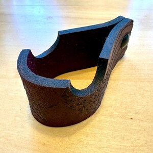 May include: A dark brown leather cuff bracelet with a textured exterior. The bracelet has a curved design with a dark green accent and a small, dark green stud. The cuff is shown on a light brown wooden surface.