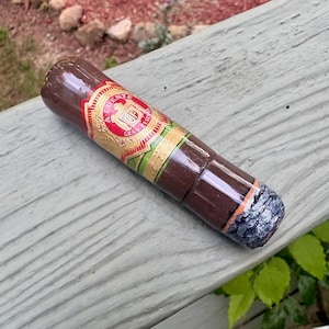 May include: A dark brown cigar with a lit, glowing grey ash at one end. The cigar features a decorative band with red, gold, and green accents, displaying the text A. FUENTE GRAN RESERVA in a circular emblem with a central crest and gold filigree.
