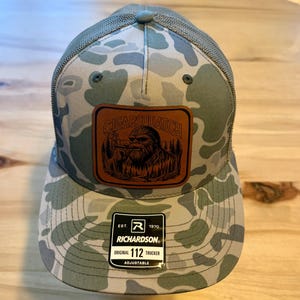 May include: A camouflage trucker hat with a brown leather patch featuring the text "CIGARSQUATCH" and an illustration of Bigfoot smoking a cigar. The hat has a mesh back and an adjustable strap.