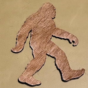 May include: A wooden cutout of Bigfoot, the legendary creature, in a walking pose. The figure is brown with a textured, hairy appearance and a black outline. The background is a muted olive green.