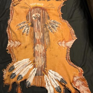 May include: Hand-painted Native American hide featuring a central design with a sun, feathers, and geometric patterns in shades of brown, white, and black. The hide has a natural, irregular shape with a rough edge.