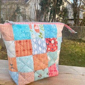 May include: A patchwork fabric pouch with a zipper closure. The pouch is made of various squares of fabric in shades of pink, blue, orange, and white. The fabric squares feature floral and geometric patterns. The zipper pull is a matching fabric loop.