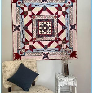 May include: A colorful quilt titled "Arabesque" designed by Sandra Walker, featuring a geometric pattern with red, pink, and blue star and square shapes. The quilt hangs on a wall above a chair and a small table.