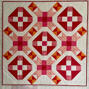 Pode incluir: Uma colcha com um design de patchwork com quadrados e losangos vermelhos, rosa e laranja. A colcha é acolchoada com fio branco e tem um fundo branco.