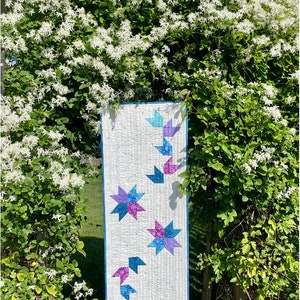 May include: A white quilted table runner with a blue border and a design of blue, pink, and purple stars. The text "Rosebud Garland" and "Designed by Sandra Walker" is at the top of the image. The text "Seasonal décor made easy with a runner and coordinating placemats" and "Precuts or scraps; beginner-friendly" is at the bottom of the image.