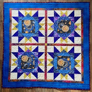 Peut inclure: Une courtepointe carrée composée de quatre blocs en forme d'étoile avec des motifs célestes, incluant des soleils, des lunes et des étoiles dans des tons de bleu, violet et or. Elle possède une bordure bleue et le texte SandraJaneQuilts est visible.