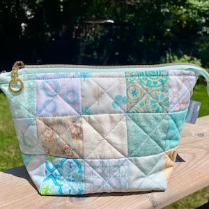 May include: A quilted zip pouch featuring a patchwork design of light blue, teal, and white fabric squares. The pouch has a gold-toned metal zip pull with a circular ring and a small white fabric tag attached to the side seam.
