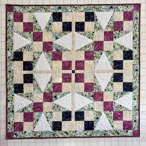 Puede incluir: Una colcha con un patrón geométrico de triángulos blancos, cuadrados burdeos y cuadrados crema sobre un fondo floral verde.