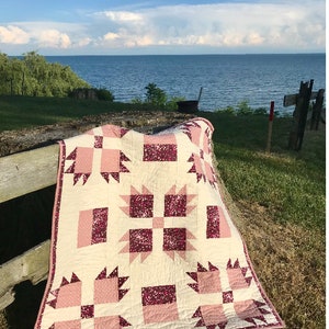 Puede incluir: Una colcha de bebé rosa y blanca con un diseño de pata de oso. La colcha mide 40 1/2 pulgadas por 44 1/2 pulgadas. La colcha está colocada en un banco de madera frente a un lago.