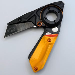 May include: A black and orange utility knife with a partially exposed blade. The knife features a black handle with orange accents and a circular design element. The blade has a geometric design etched on it.