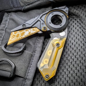 Puede incluir: Una navaja plegable con un diseño negro y dorado. La hoja presenta un patrón de panal y la palabra "HYDRA". El mango tiene una sección transparente teñida de amarillo. El cuchillo se muestra sobre una superficie negra texturizada.