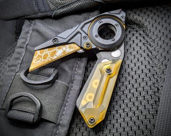 Può includere: Un coltello multiuso pieghevole con un design nero e oro. La lama presenta un motivo a nido d'ape e la scritta "HYDRA". L'impugnatura ha una sezione trasparente colorata di giallo. Il coltello è mostrato su una superficie nera strutturata.