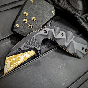 May include: A black and gold tactical knife with a geometric honeycomb pattern on the blade. The knife has a textured handle and is shown with a black sheath. The blade is angled and appears sharp.