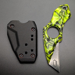 May include: A small, tactical knife with a neon green and black marbled handle and a silver blade. The knife has a finger ring and is accompanied by a black sheath. The blade features a geometric design. The knife and sheath are on a dark gray surface.