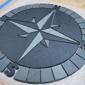 May include: A circular, gray compass rose design with a central star and directional letters. The compass rose is made of dark gray material with lighter gray accents and is set on a white mesh surface.