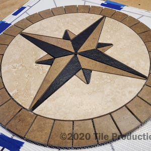 May include: Circular tile mosaic featuring a compass rose design. The compass rose has a black and tan star shape in the center, surrounded by a beige background and a brown border. The mosaic is on a white mesh.