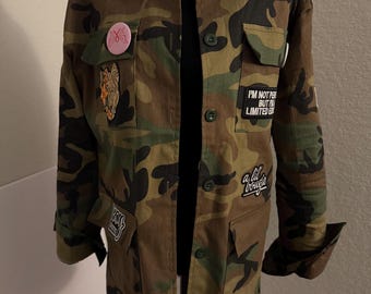 May include: Camouflage print jacket with multiple patches. The jacket features a button-up front, multiple pockets, and patches with text and floral designs. The jacket is displayed on a mannequin.