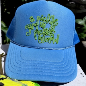 May include: A light blue trucker hat featuring a foam front panel and mesh back. The front panel displays embroidered lime green text that reads, "a pickle girl in a pickles world," written in a stylized, rounded, and bubbly cursive font.