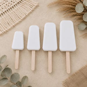 Pode incluir: Quatro objetos em forma de picolé de tamanhos variados, cada um apresentando um topo retangular arredondado e liso preso a um palito de madeira marrom claro. Os objetos estão dispostos em uma fileira do menor para o maior, com acabamento branco.