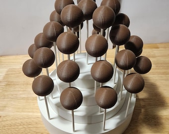 May include: A tiered cake stand with 24 chocolate cake pops arranged in a circular pattern. The cake pops are brown and have a textured surface. The cake stand is white and has three tiers.
