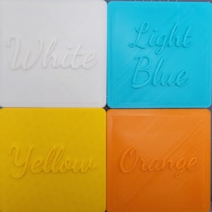 Puede incluir: Cuatro posavasos cuadrados en blanco, azul claro, amarillo y naranja. Cada posavasos presenta letras en relieve en cursiva que identifican su color: White, Light Blue, Yellow y Orange. Las superficies muestran una sutil textura diagonal.