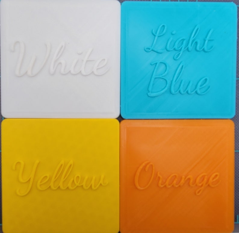 Puede incluir: Cuatro posavasos cuadrados en blanco, azul claro, amarillo y naranja. Cada posavasos presenta letras en relieve en cursiva que identifican su color: White, Light Blue, Yellow y Orange. Las superficies muestran una sutil textura diagonal.