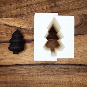 以下が含まれることがあります： クリスマスツリーの形をしたオブジェクトを作るための白いシリコンモールド。モールドは開いており、モールドから作られた黒いクリスマスツリーが隣にあります。