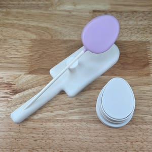 May include: A white plastic cake decorating tool set. The set includes a light purple oval-shaped lollipop, a white oval-shaped cutter, and a white handle with a flat base. The tools are on a wooden surface.