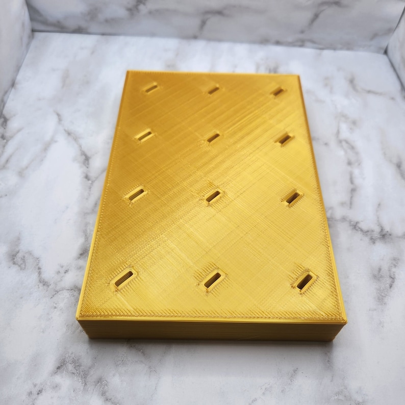 Puede incluir: Un objeto rectangular impreso en 3D de color dorado con una superficie superior plana que presenta una cuadr&iacute;cula de doce peque&ntilde;as aberturas rectangulares espaciadas uniformemente. La superficie tiene un patr&oacute;n de textura diagonal.