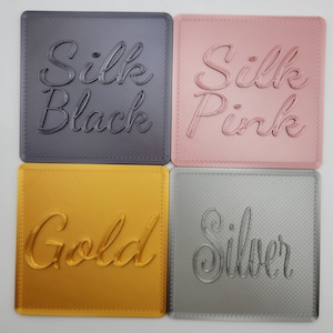 Puede incluir: Cuatro posavasos cuadrados con un acabado texturizado en diagonal. Arriba a la izquierda, gris oscuro con el texto Silk Black. Arriba a la derecha, rosa claro con el texto Silk Pink. Abajo a la izquierda, dorado con el texto Gold. Abajo a la derecha, plateado con el texto Silver.