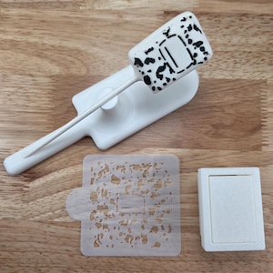 May include: A white cake decorating tool set. The set includes a spatula with a rectangular head featuring black spots and a central design, a stencil, and a small rectangular container.