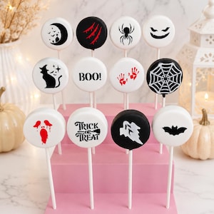 May include: A collection of twelve round Halloween-themed cake pops arranged on a pink tiered stand. The pops feature white or black icing with various designs including a crescent moon, red claw marks, a spider, a ghost face, a cat, the text BOO!, red handprints, a spiderweb, a dripping red face, the text TRICK OR TREAT, a white ghost, and a black bat.