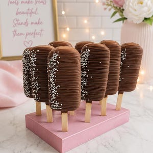 Puede incluir: Un conjunto de siete cake pops cubiertos de chocolate sobre palitos de madera, dispuestos en un soporte rectangular de madera rosa. Cada cake pop est&aacute; ba&ntilde;ado en chocolate con leche con un patr&oacute;n de l&iacute;neas y perlas de az&uacute;car blancas.