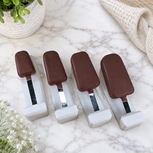 Puede incluir: Cuatro objetos rectangulares de color chocolate con forma de polo helado sobre bases de plástico blanco. Cada pieza presenta una parte superior marrón oscuro que recuerda a un helado en palo, montada sobre un soporte rectangular blanco.