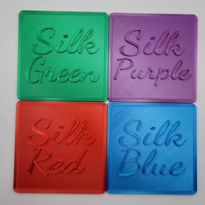 Puede incluir: Cuatro posavasos cuadrados con acabado brillante, cada uno con texto en cursiva en relieve. El posavasos verde dice Silk Green, el morado Silk Purple, el rojo Silk Red y el azul Silk Blue. Cada posavasos tiene una textura diagonal.