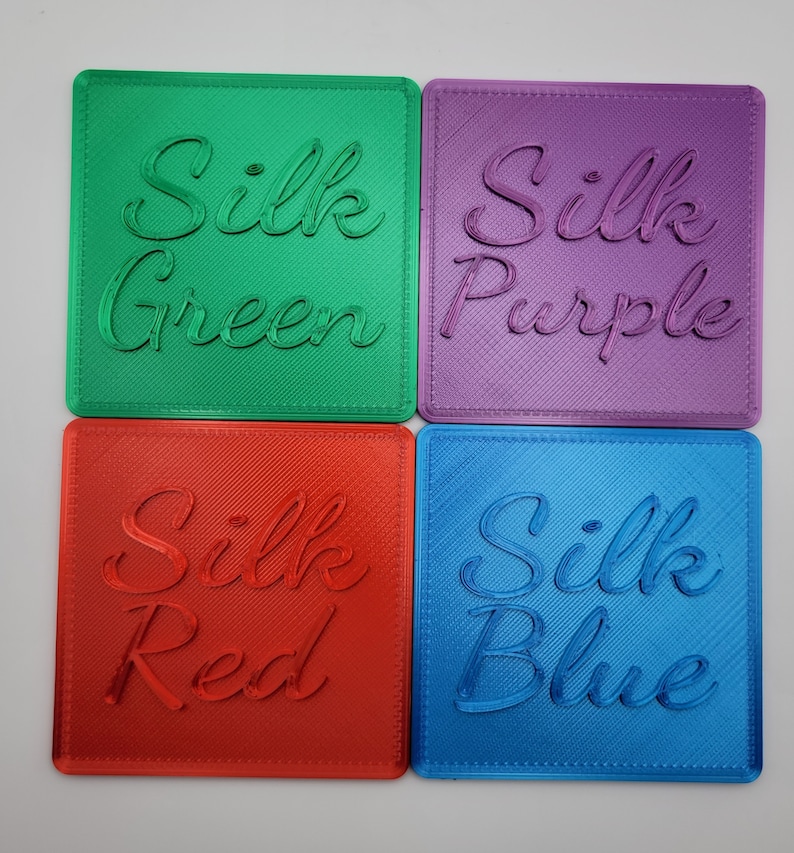 Puede incluir: Cuatro posavasos cuadrados con acabado brillante, cada uno con texto en cursiva en relieve. El posavasos verde dice Silk Green, el morado Silk Purple, el rojo Silk Red y el azul Silk Blue. Cada posavasos tiene una textura diagonal.