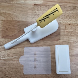 May include: A white and yellow measuring tool with a ruler-like design. The tool is composed of a yellow rectangular piece with black markings, a white handle, and a white base. Also included are a clear measuring guide and a white rectangular piece.