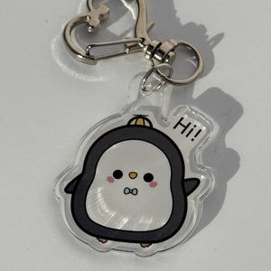 以下が含まれることがあります： 透明なアクリルキーホルダー。黒と白の体、ピンクの頬、小さな青い蝶ネクタイをしたペンギンのキャラクターが描かれています。ペンギンは黄色い帽子をかぶって手を振っており、横には黒い文字で「Hi!」と書かれています。銀色のハート型の留め具が付いています。
