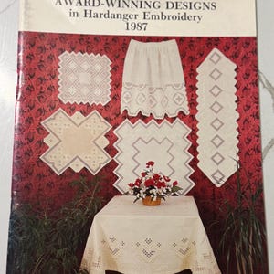 May include: A softcover book titled AWARD-WINNING DESIGNS in Hardanger Embroidery 1987, compiled by Susan L. Meier and Rosalyn K. Watnemo. The cover features white Hardanger embroidery pieces displayed against a dark red floral patterned background.