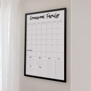 May include: A framed, white monthly calendar with a black border. The calendar grid is blank, with the days of the week across the top. The words "Greenwood Family" are written in cursive at the top. Below the calendar are sections for notes and a to-do list.