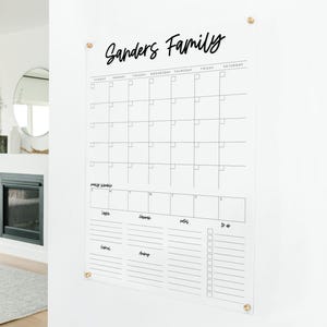 May include: Clear acrylic family calendar with the name "Sanders Family" in black script. The calendar features a monthly grid, a section for weekly schedules, and a to-do list. It is mounted on a white wall with gold-colored hardware.