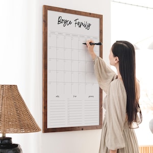 May include: A large, framed whiteboard calendar with the text "Boyce Family" at the top. A person is writing on the calendar with a black marker. The calendar has a grid layout for the days of the week and space for notes.