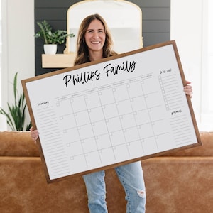May include: A large, rectangular wooden-framed whiteboard calendar with the text "Phillips Family" in a handwritten font. The calendar has a grid for the days of the month, a to-do list, and space for notes. The person holding the calendar is wearing blue jeans.