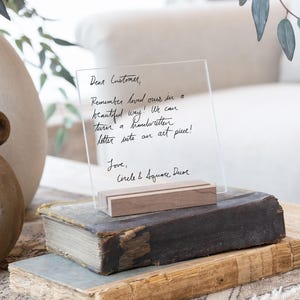 This is a personalized acrylic sign in a wood stand. Handwritten letter written in black text. The sign is sitting on a side table on top of decor books.