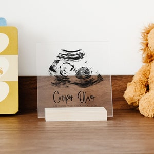 This clear acrylic sign announces baby's birth. Ultrasound, name, birth, time, and date are in black. The square sign is in a blonde wood stand sitting on a dresser.