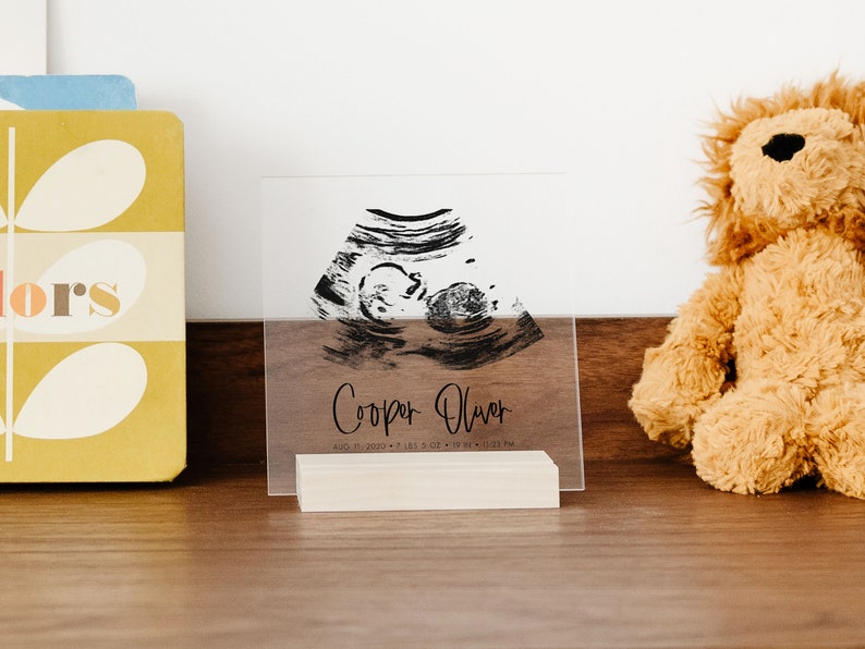 This clear acrylic sign announces baby's birth. Ultrasound, name, birth, time, and date are in black. The square sign is in a blonde wood stand sitting on a dresser.