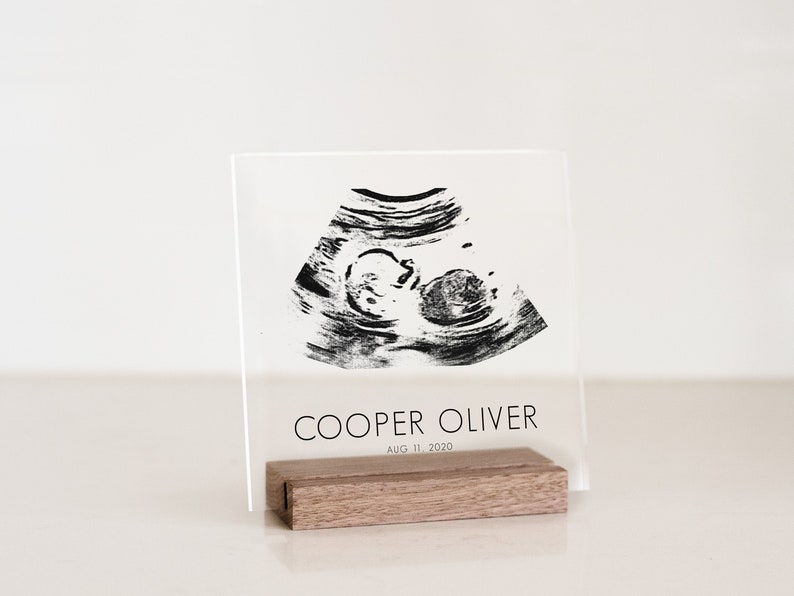 This baby announcement sign is made with clear square acrylic. The baby ultrasound and birthdate are in black print. The sign is in a walnut wood stand on a table.