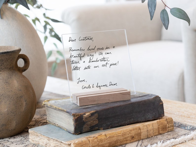 This is a personalized acrylic sign in a wood stand. Handwritten letter written in black text. The sign is sitting on a side table on top of decor books.