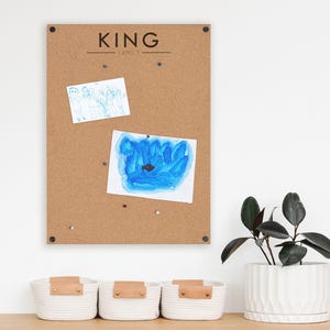 May include: A corkboard with the text "KING FAMILY" and two drawings pinned to it. Below are three white woven baskets with leather accents. A potted plant with dark green leaves sits to the right.
